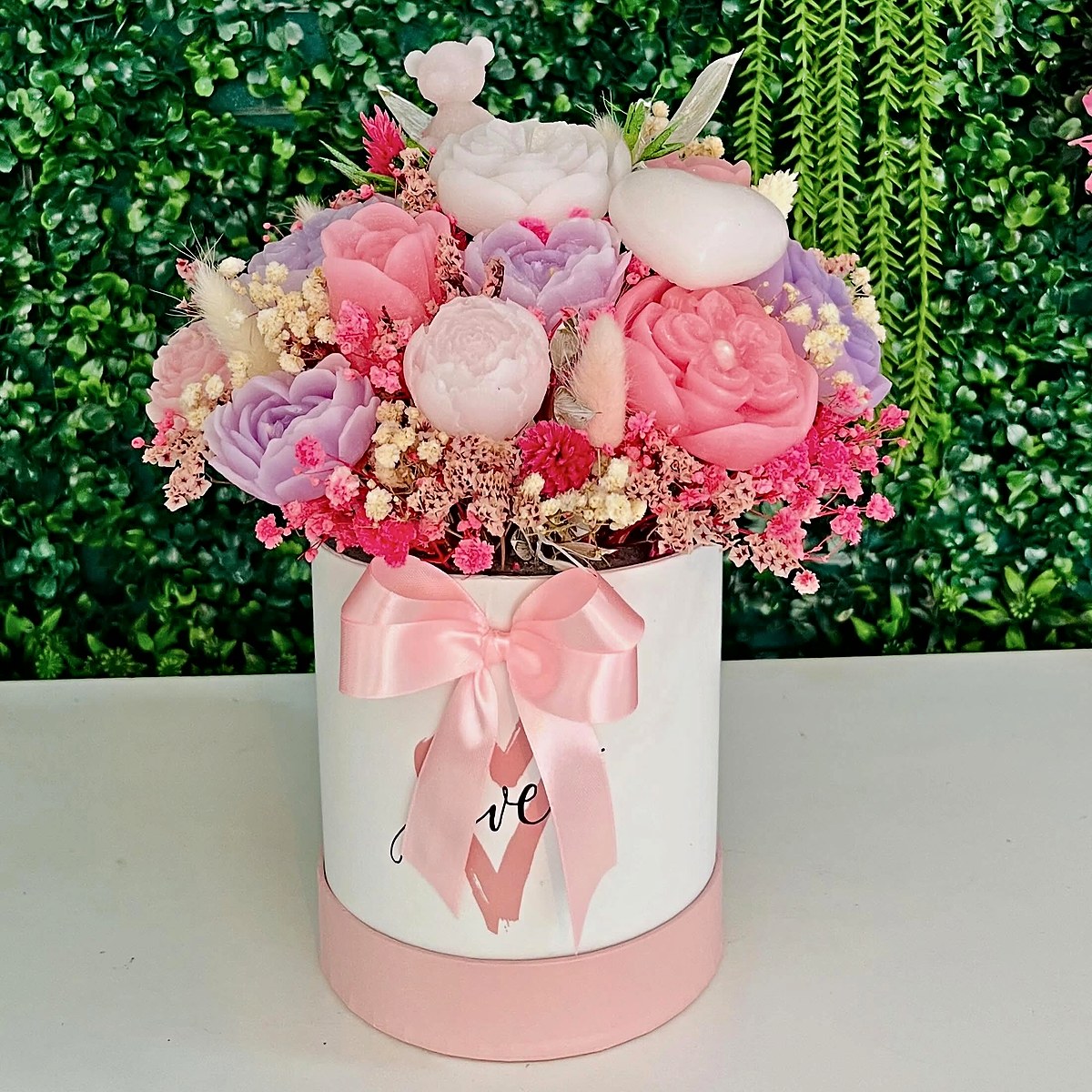 Caixa de flores em tons de rosa e lilás com flores secas, ursinho e laço cor-de-rosa, prenda preparada pela Florabella em Fafe