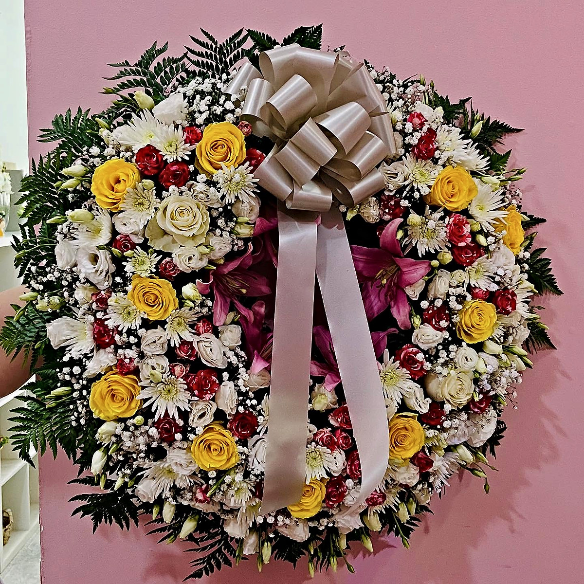 Coroa de flores para funeral com rosas amarelas e brancas, mini rosas vermelhas, lisianthus, crisântemos, lírios cor-de-rosa e laço prateado, feita pela Florabella em Fafe