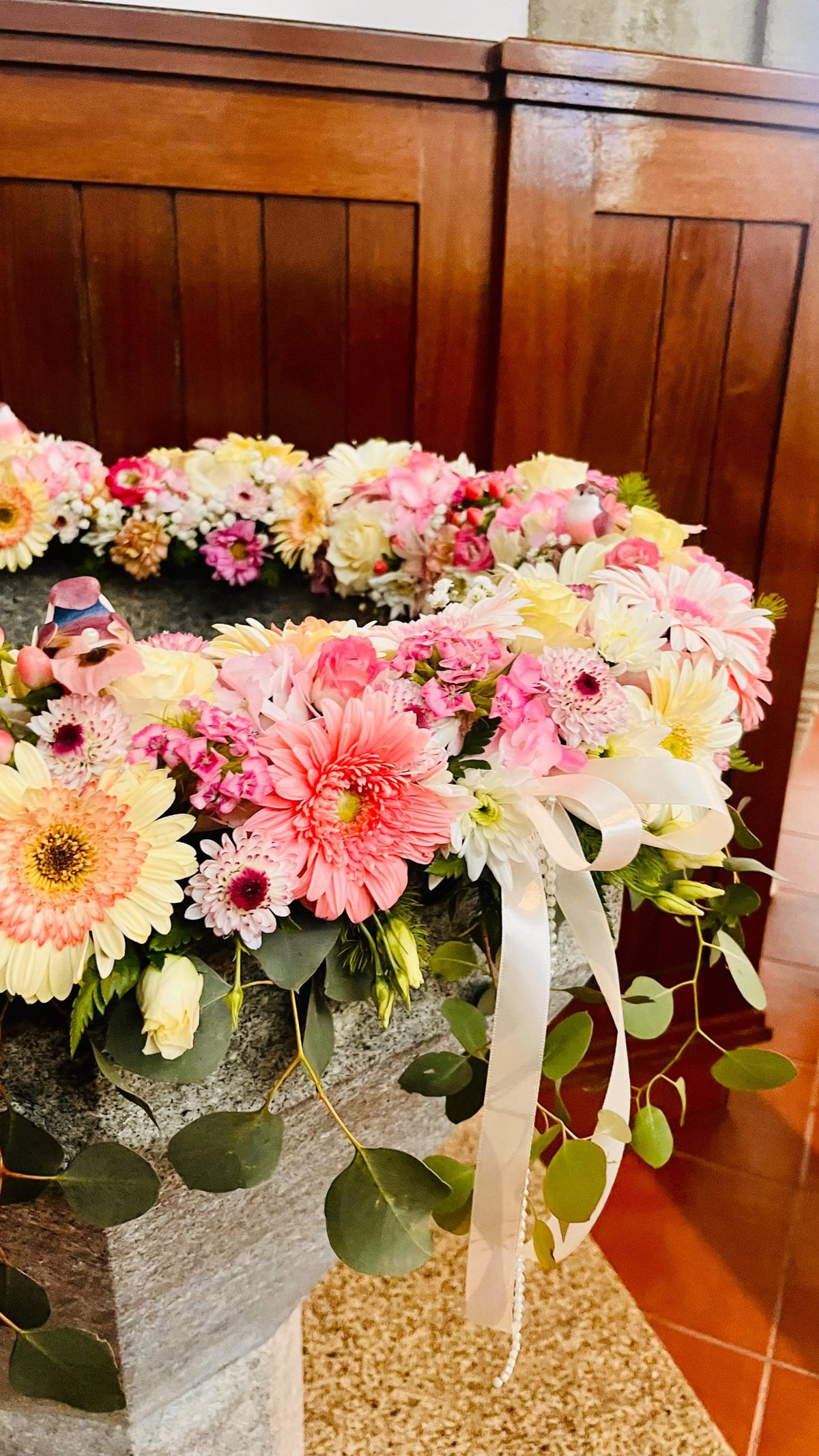 Coroa de flores para pia batismal com gérberas, cravinas, laço branco com pérolas e eucalipto, pela Florabella em Fafe