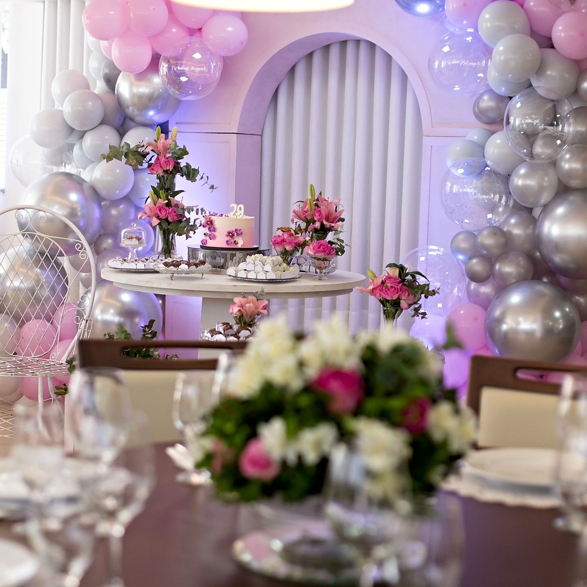Decoração de festa de aniversário com arco de balões cor-de-rosa e prateados, mesa do bolo com lírios e rosas cor-de-rosa e centro de mesa com flores, pela Florabella em Fafe
