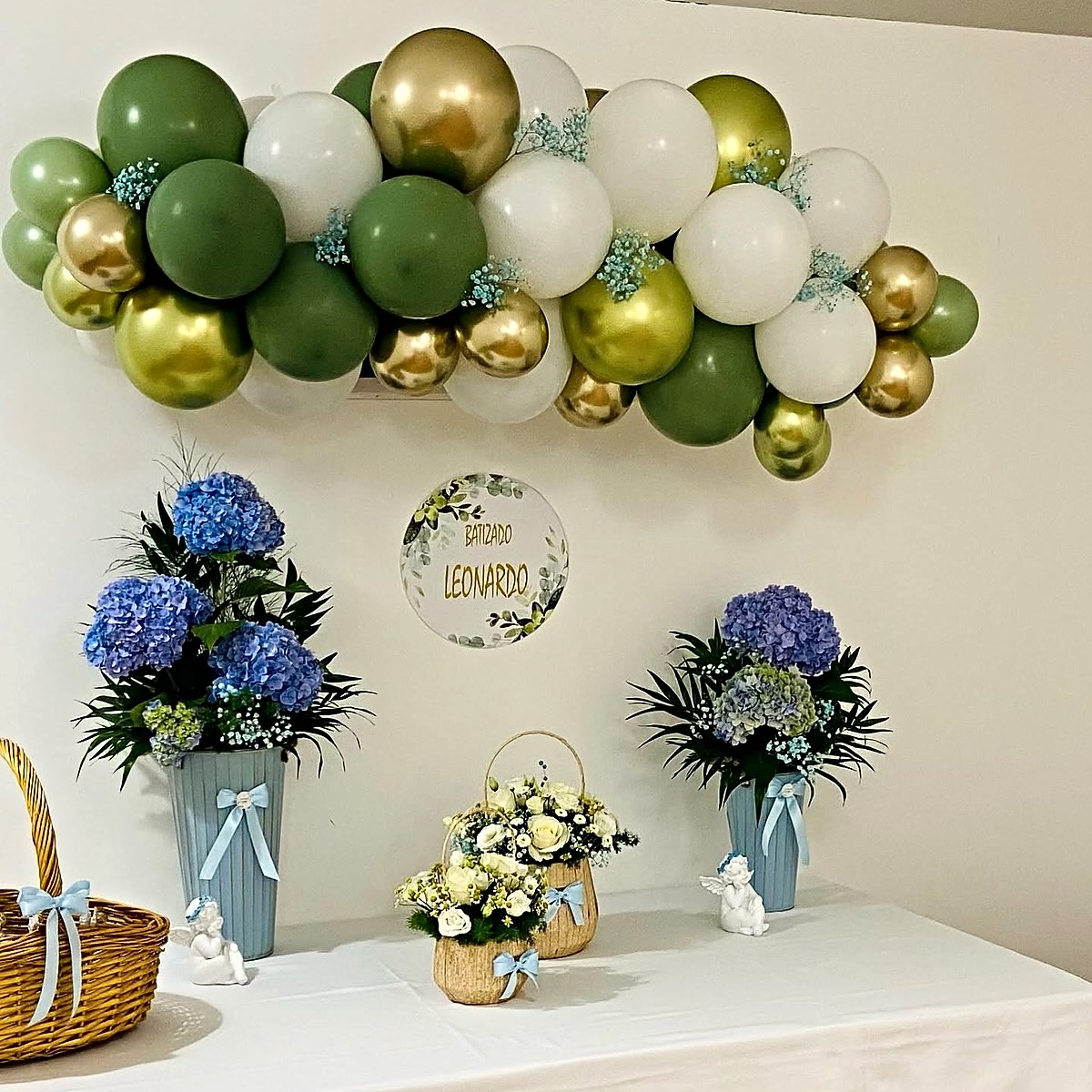 Decoração de festa de batizado com arco de balões verdes, brancos e dourados, arranjos de hortênsias azuis e cestos de rosas brancas, pela Florabella em Fafe