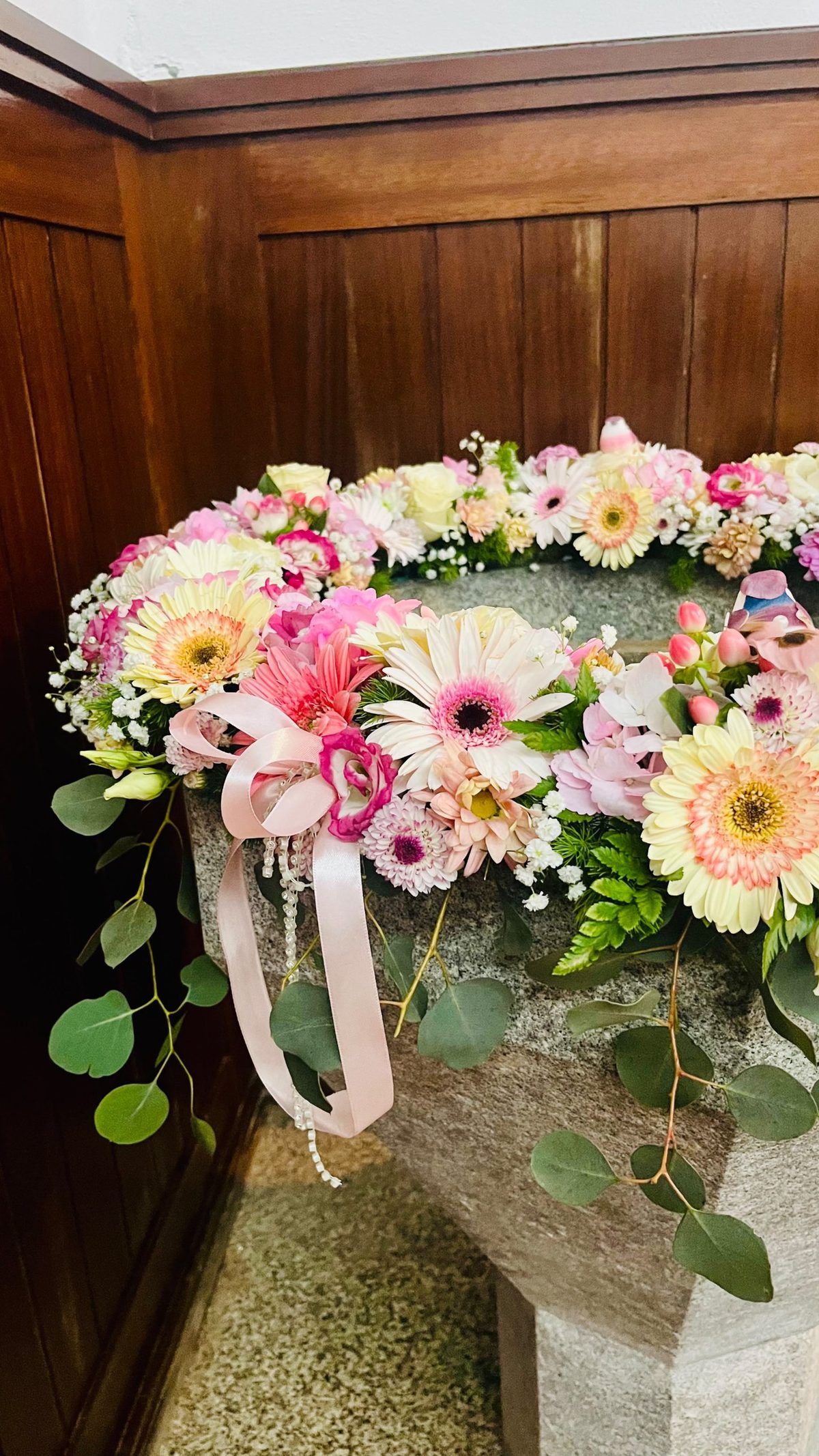 Pormenor da decoração da pia batismal com gérberas, crisântemos, lisianthus, laço cor-de-rosa e eucalipto, pela Florabella em Fafe