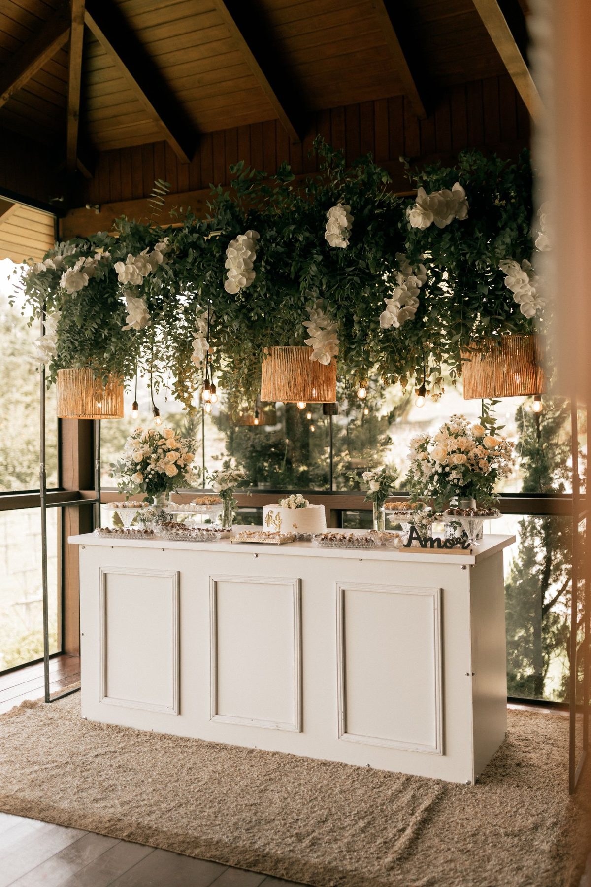 Mesa de doces de casamento com decoração suspensa de folhagem e orquídeas brancas e arranjos de rosas brancas, pela Florabella em Fafe