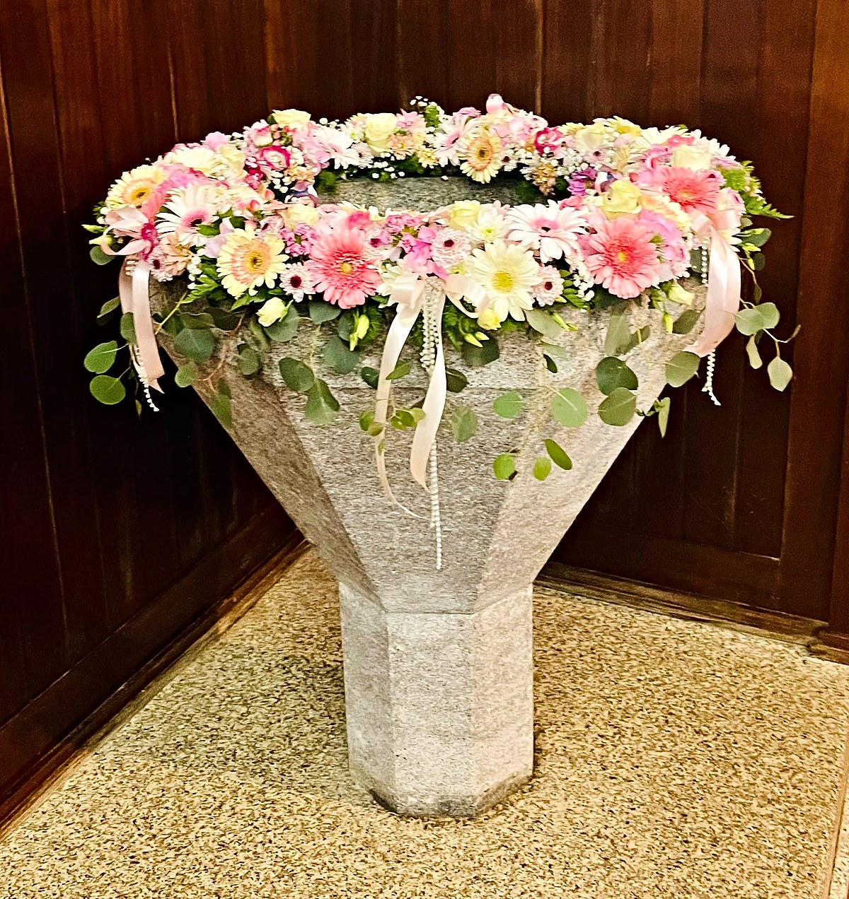 Pia batismal de granito decorada com uma coroa de gérberas, rosas, cravos e eucalipto, com laços e fios de pérolas, para batizado, pela Florabella em Fafe